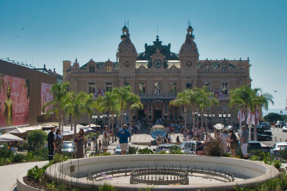 Un grande edificio decorato si erge dietro una piazza affollata con palme, una fontana circolare e molte persone sotto un cielo sereno.