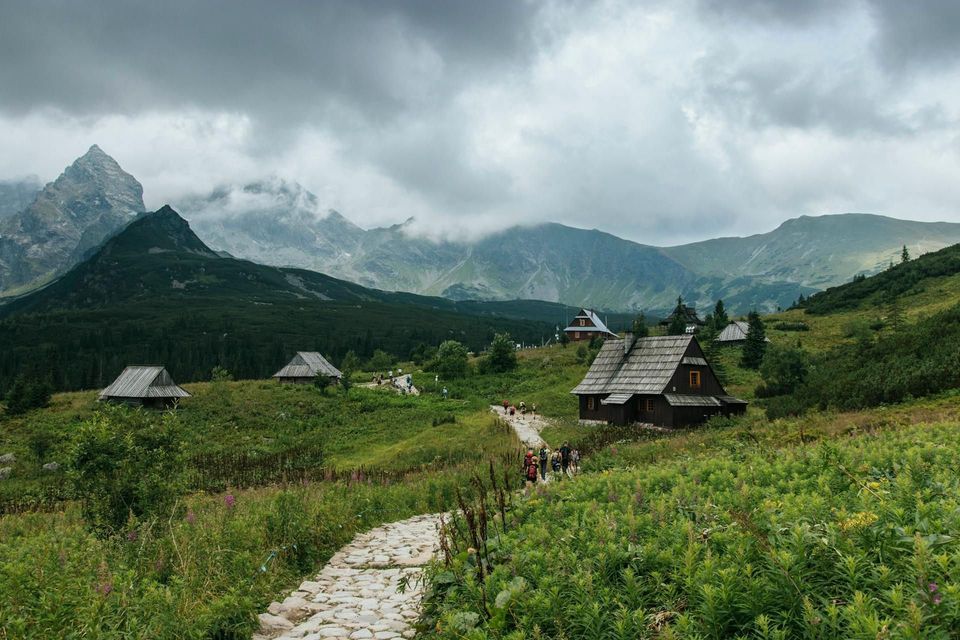 Eine WeRoad-Gruppenreise führt auf einem Kopfsteinpflasterweg durch ein grünes Bergtal mit Holzhütten unter bewölktem Himmel.