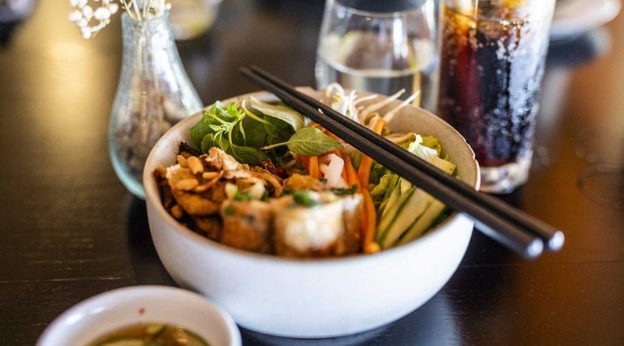 Primo piano di una ciotola bianca di insalata di noodle asiatica con bacchette, servita su un tavolo scuro con un bicchiere di bibita sullo sfondo.