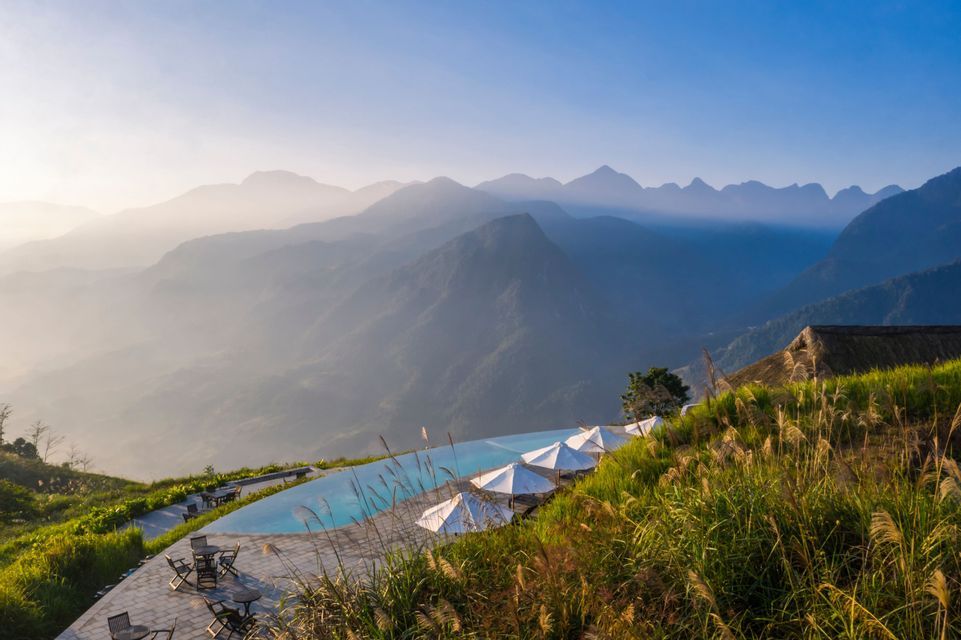 Una piscina a sfioro su una collina erbosa con tavoli e ombrelloni si affaccia su una vasta catena montuosa velata all'alba.