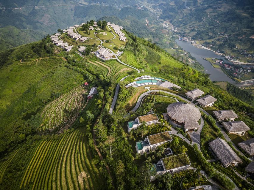 Una vista aerea di un resort con ville e piscine su una rigogliosa montagna verde, che si affaccia su campi terrazzati e una valle fluviale.