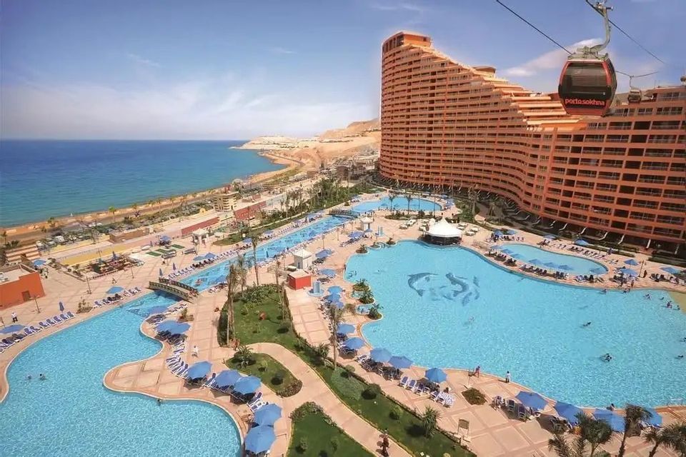 Una vista aérea de un gran complejo turístico junto al mar con amplias piscinas, un hotel curvo y un teleférico que pasa por encima.