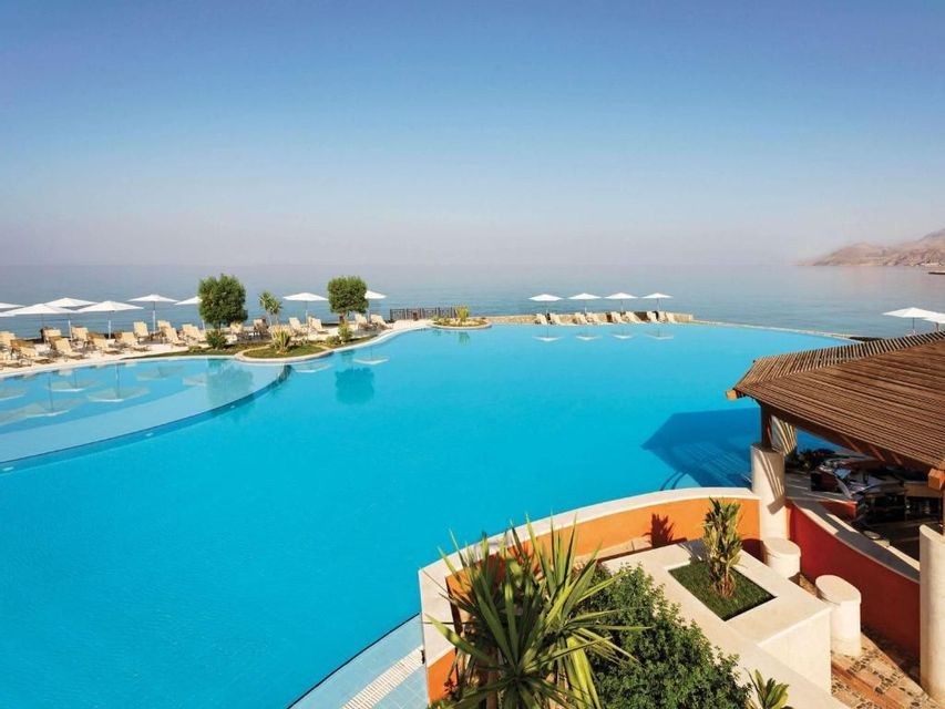 Una gran piscina infinita en un complejo turístico con vistas al mar, rodeada de tumbonas y sombrillas blancas bajo un cielo despejado.