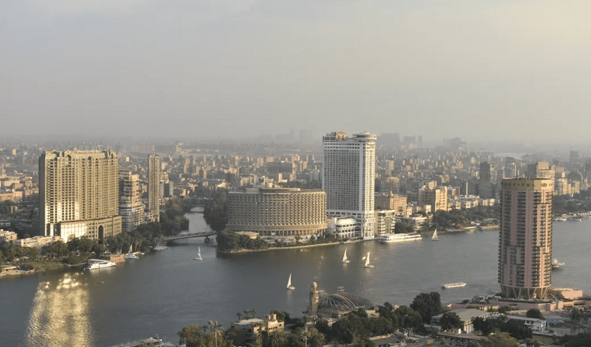 Una vista aérea de una ciudad en expansión con veleros en un río amplio que fluye entre altos edificios bajo un cielo brumoso.