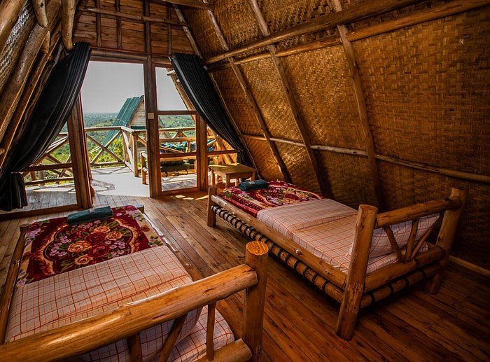 El interior de una cabaña rústica tipo A con dos camas de madera, paredes tejidas y una puerta abierta que da a un balcón y colinas verdes.