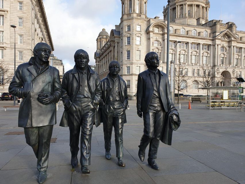 Quattro statue in bronzo a grandezza naturale dei Beatles che camminano su una piazza pavimentata di fronte a grandi edifici storici.