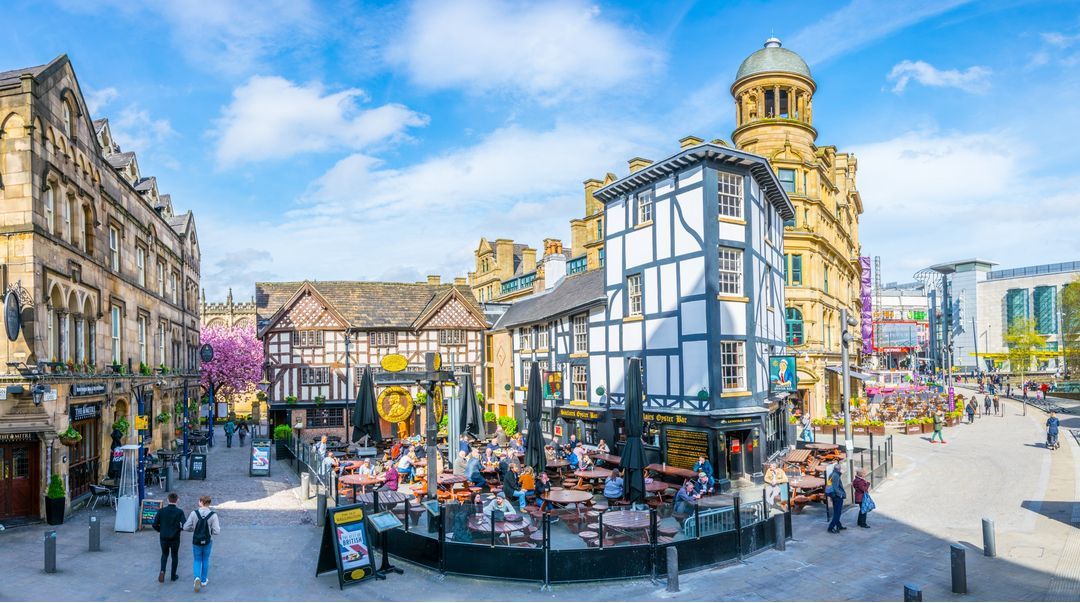 Una vivace piazza cittadina con persone ai tavoli all'aperto, circondata da edifici storici in stile Tudor e in pietra sotto un cielo blu.
