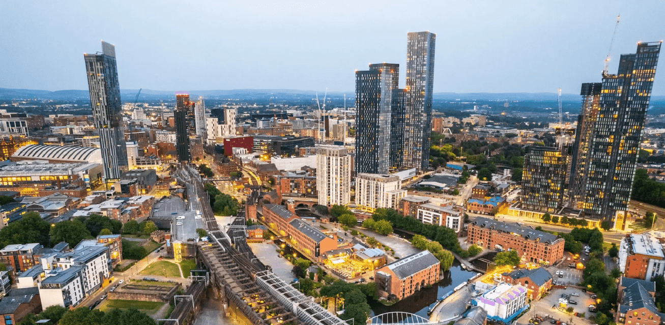Una vista aerea dello skyline di una città al tramonto, con un mix di grattacieli moderni ed edifici in mattoni più antichi lungo canali e binari ferroviari.