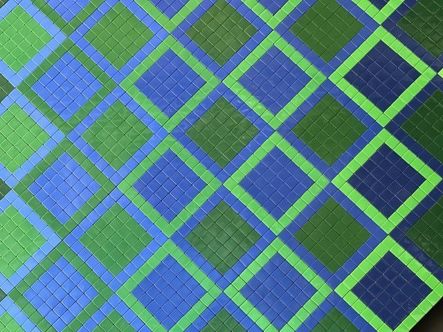 Un primo piano di un mosaico composto da piccole tessere quadrate che formano un motivo a quadri diagonale nelle tonalità del verde e del blu.