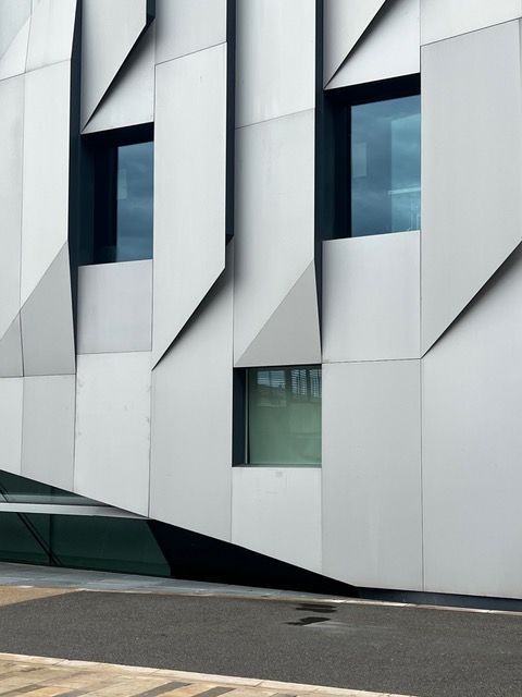 La facciata di un edificio moderno con pannelli angolari, geometrici, metallici e diverse finestre scure.