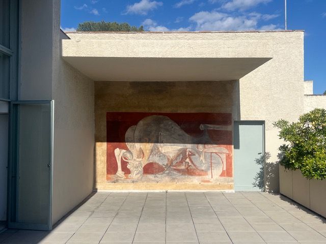 Un murale astratto nei toni del rosso e del grigio sulla parete esterna del patio di un edificio moderno sotto un cielo blu.