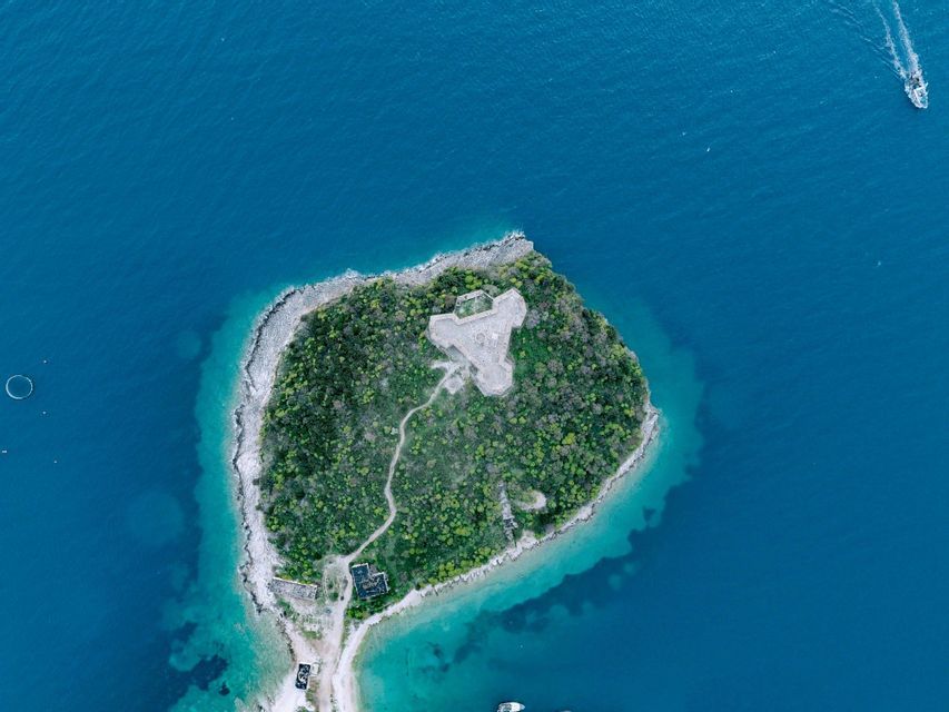 Vista aérea cenital de una pequeña isla en forma de corazón con un fuerte de piedra rodeado de mar turquesa y azul profundo.