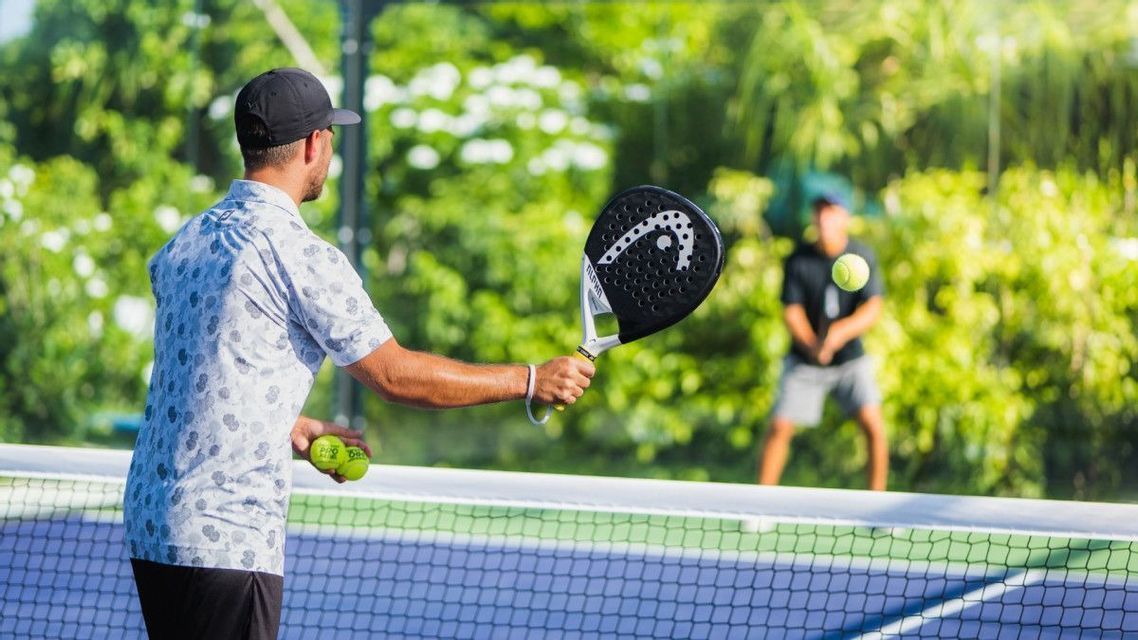 Un uomo visto di spalle serve con una racchetta da padel su un campo all'aperto, con un altro giocatore sullo sfondo.