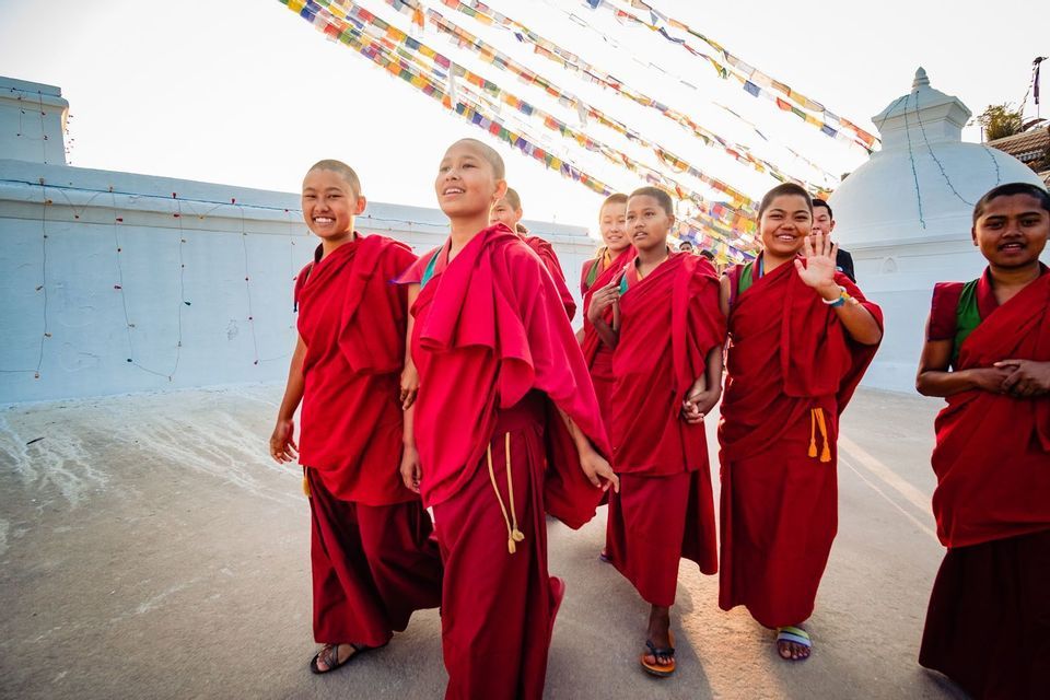 A group of young Buddhist nuns in red robes smile and walk past a white stupa under strings of colorful prayer flags.