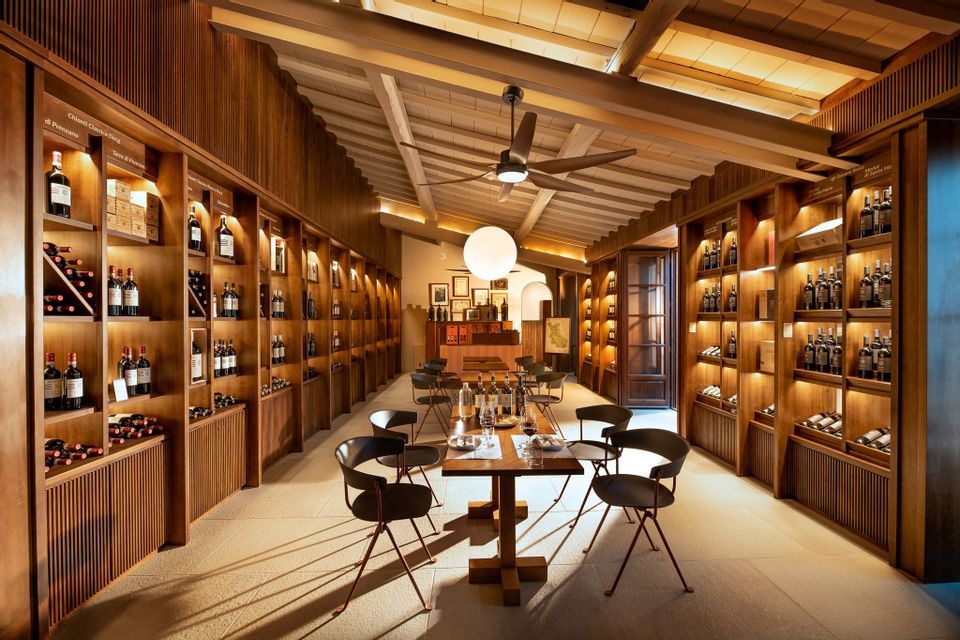 Una elegante sala degustazione vini con tavoli e sedie, circondata da scaffali in legno illuminati che espongono numerose bottiglie di vino.