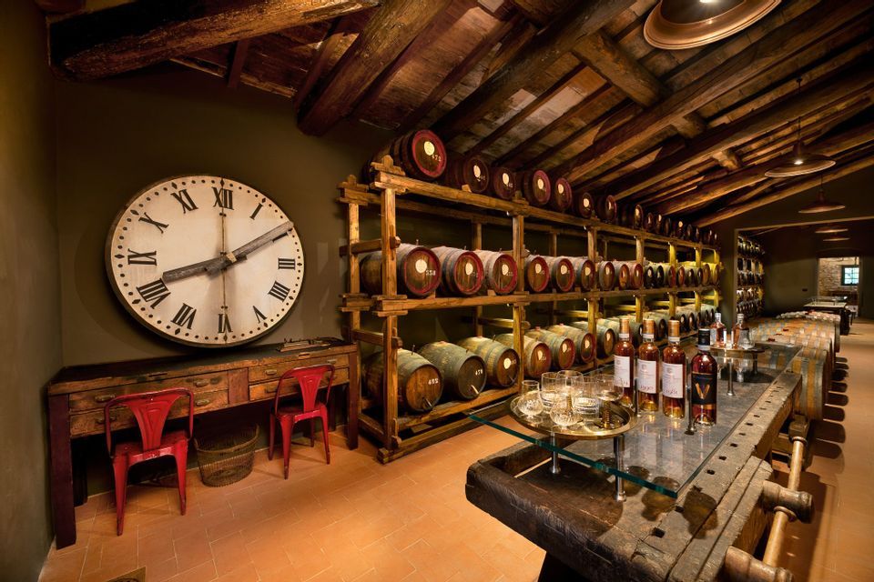 Una cantina rustica con botti di legno su scaffalature, un grande orologio a muro e un tavolo preparato con bottiglie e bicchieri per una degustazione.