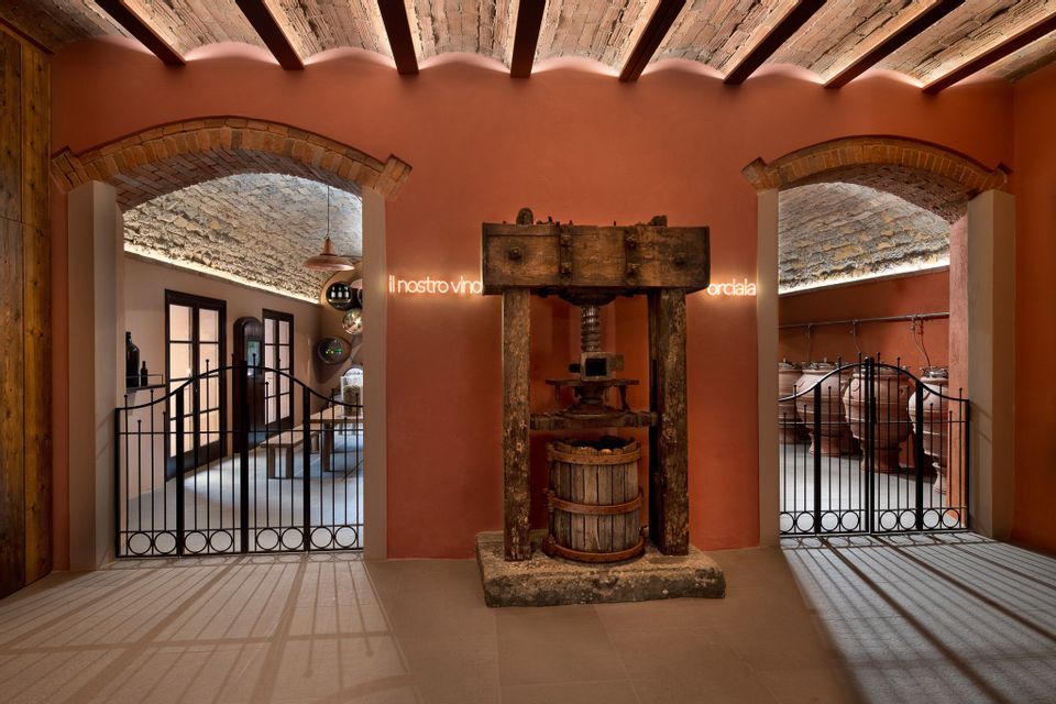 Un antico torchio vinario in legno si trova in una cantina con pareti in terracotta, affiancato da due portali ad arco con cancelli in ferro battuto.