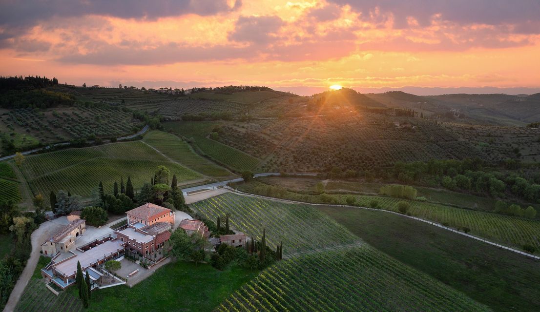 Una vista aerea di una villa con tetto in terracotta circondata da vigneti su dolci colline al tramonto.