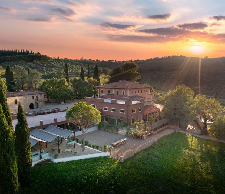 Una vista aerea di una grande villa in terracotta circondata da verdi colline e cipressi durante un tramonto colorato.