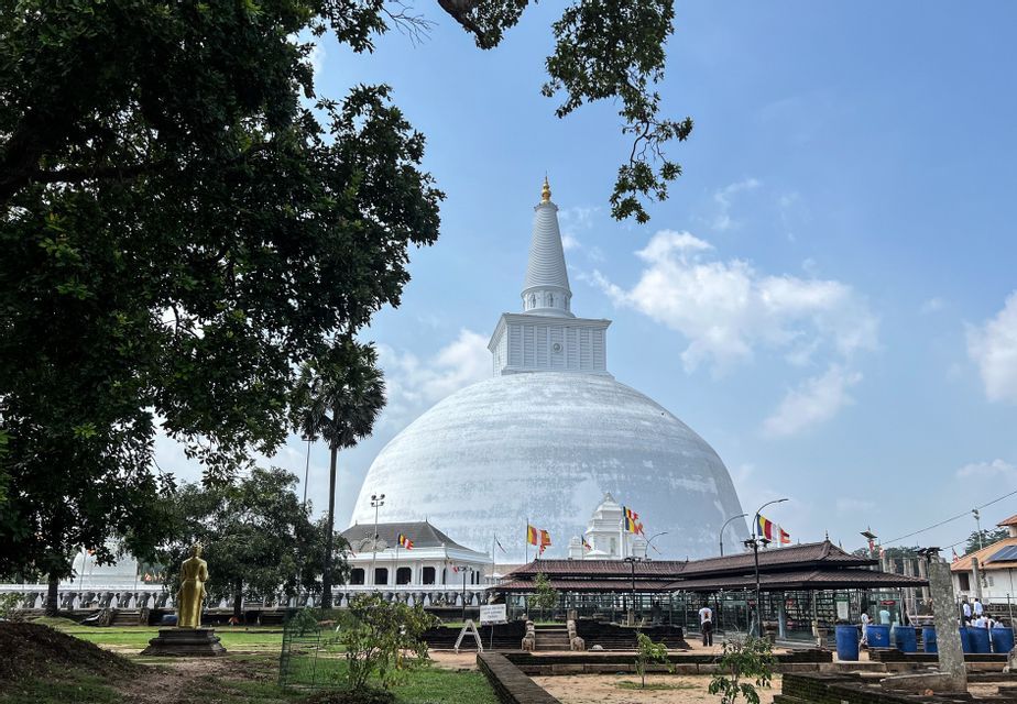 Un grande stupa bianco con una guglia dorata si erge in un complesso templare, incorniciato da alberi verdi lussureggianti sotto un cielo parzialmente nuvoloso.