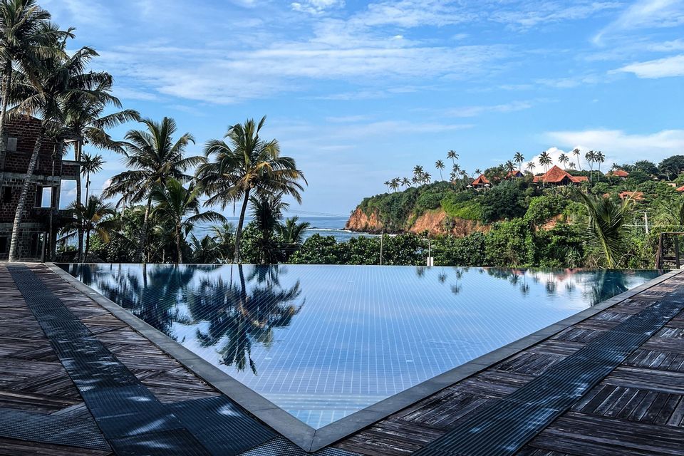 Ein Infinity-Pool mit einem Holzdeck überblickt eine tropische Küste mit Palmen, einer grünen Klippe und dem Ozean.