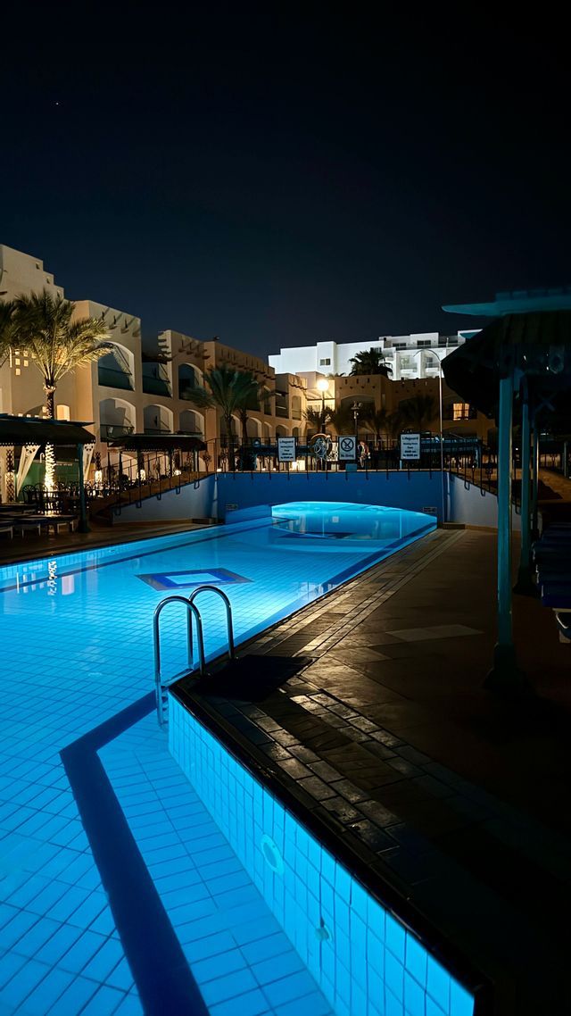 Una piscina di un resort illuminata da luci blu intense di notte, con un ponte che attraversa l'acqua.