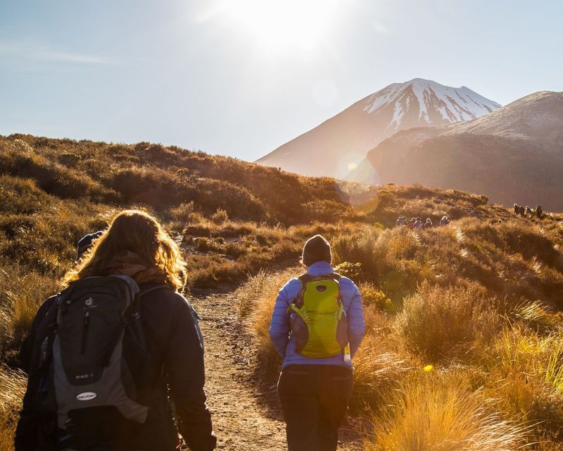 Two hikers from a WeRoad group trip walk on a sunlit trail towards a distant snow-capped mountain with others ahead.