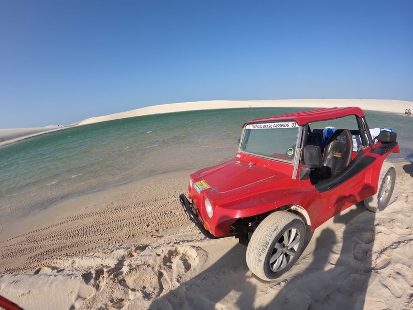 Un dune buggy rosso è parcheggiato su una spiaggia sabbiosa accanto a una laguna, con dune di sabbia bianca sullo sfondo sotto un cielo sereno.