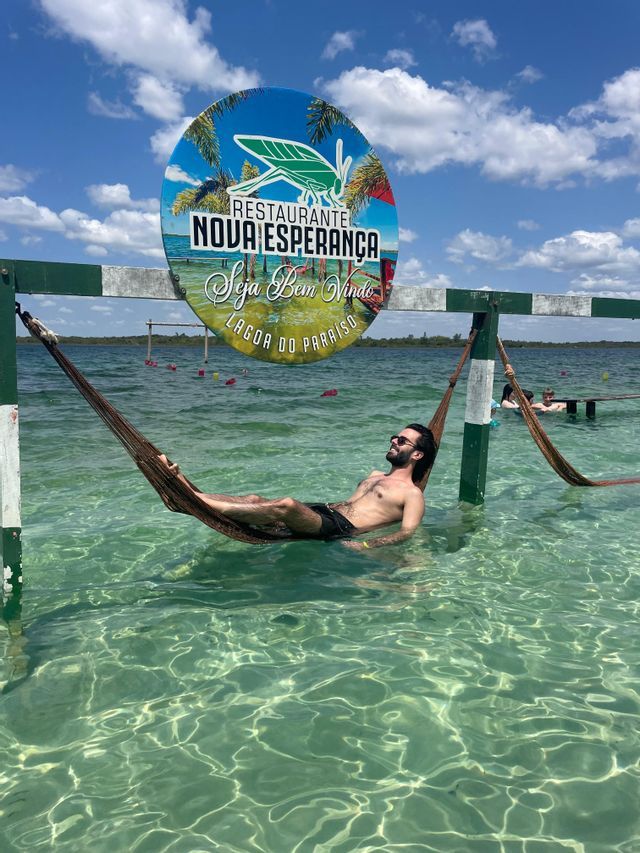 Un uomo con occhiali da sole si rilassa su un'amaca sospesa su acque turchesi limpide e poco profonde sotto un cielo azzurro con nuvole.