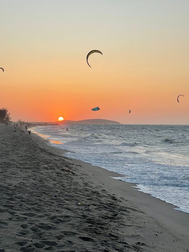Kitesurfer cavalcano le onde dell'oceano su una spiaggia sabbiosa durante un vivace tramonto arancione.