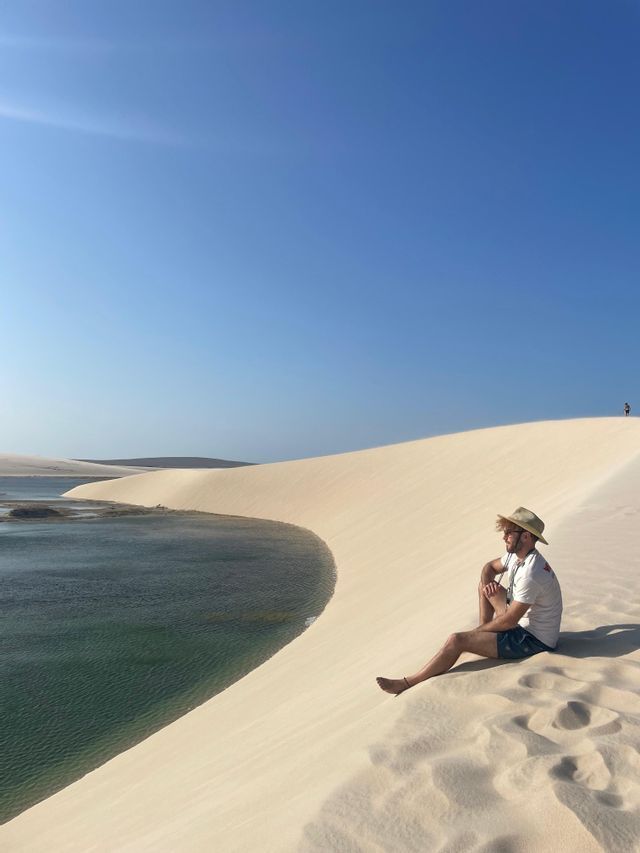 Un uomo con un cappello siede su un'enorme duna di sabbia bianca accanto a una laguna turchese sotto un cielo azzurro e limpido.