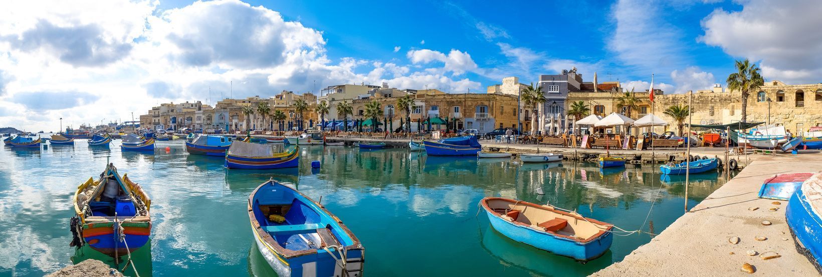 Una vista panorámica de coloridos barcos de pesca amarrados en un puerto tranquilo, con un pueblo costero y palmeras bajo un cielo parcialmente nublado.