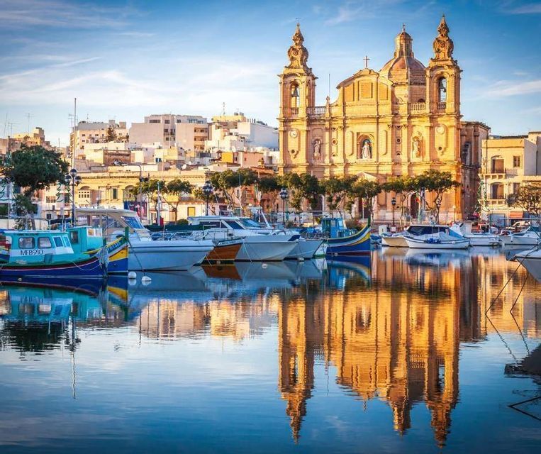 Una iglesia ornamentada con torres gemelas se refleja en las aguas tranquilas de un puerto lleno de barcos atracados.