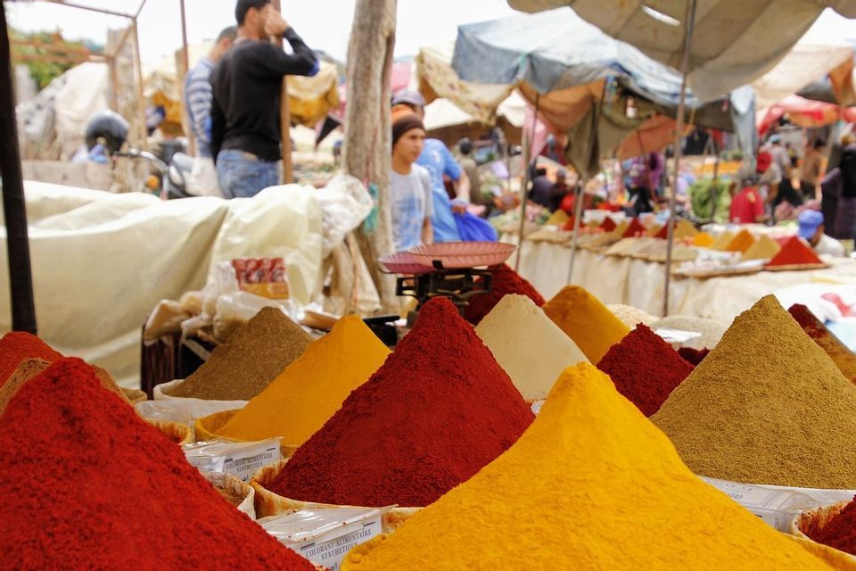 Un primo piano di varie spezie colorate disposte in mucchi conici in un mercato all'aperto, con persone sfocate sullo sfondo.