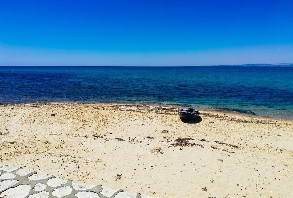 A small dark boat is beached on a sandy shore in front of a calm, blue sea under a cloudless sky.