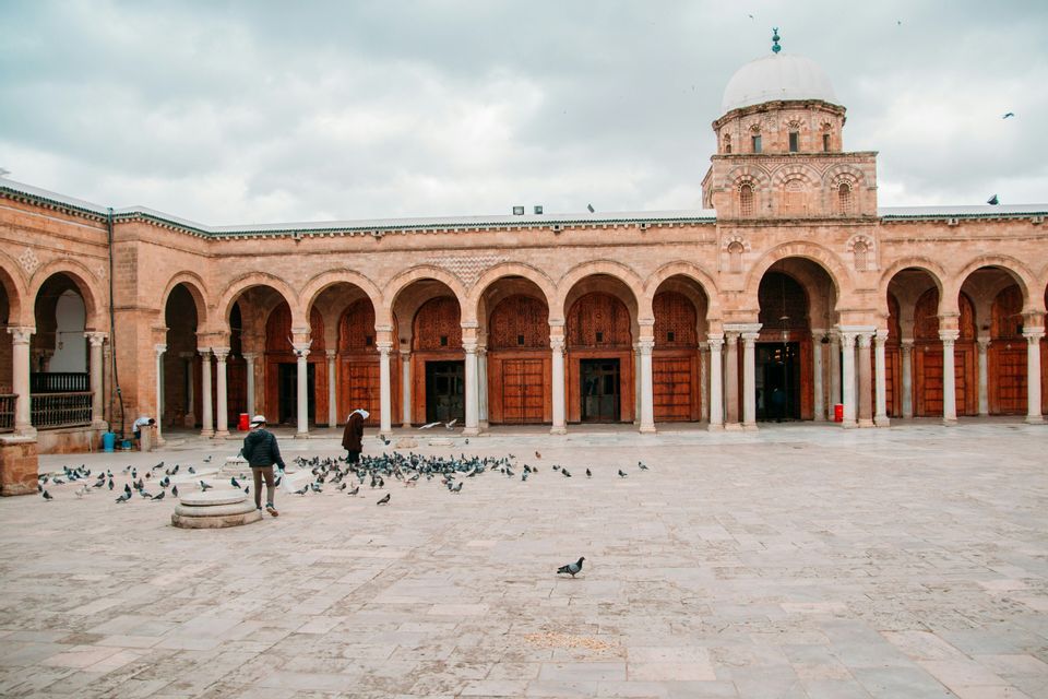 Two people walk in a large stone courtyard surrounded by an arched colonnade as pigeons gather on the ground.