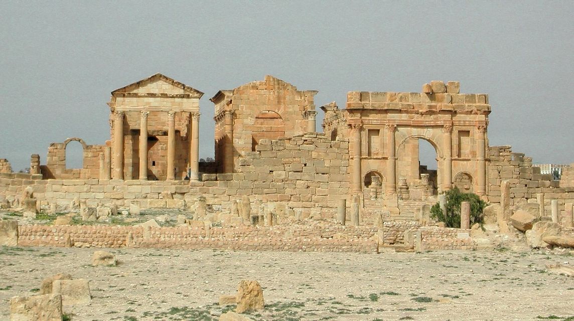 Ancient stone ruins, including a temple and an arch, stand in a vast, arid landscape under a hazy sky.
