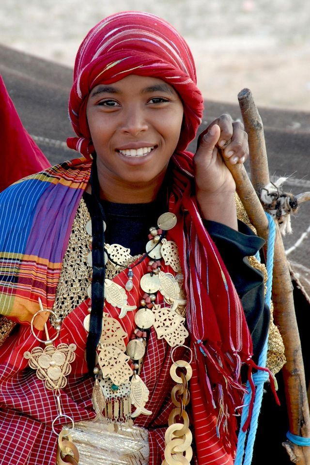 A young person smiles while wearing a red headwrap, a colorful shawl, and traditional metallic necklaces.