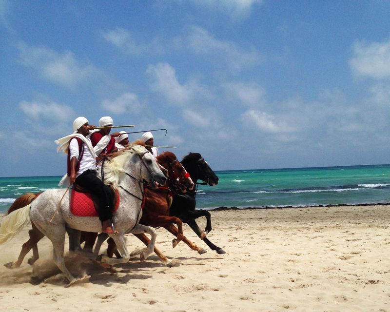 Vier Männer in traditioneller Kleidung galoppieren auf Pferden an einem Sandstrand am türkisfarbenen Meer unter blauem Himmel.