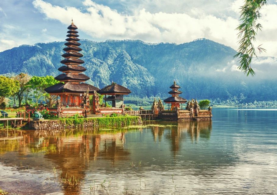 Un complejo de templos estilo pagoda a orillas de un lago, con reflejos en el agua y grandes montañas al fondo.