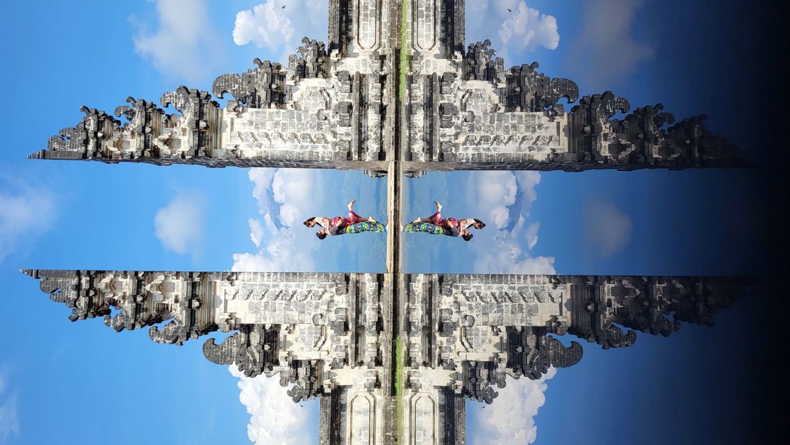 Un reflejo caleidoscópico de una persona recostada sobre una superficie colorida entre portones de piedra ornamentados bajo un cielo azul y nublado.