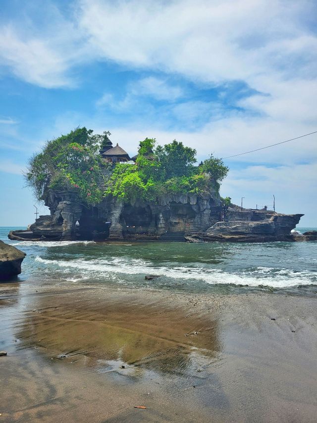 Un templo se asienta sobre un islote rocoso cubierto de árboles en el mar, con suaves olas lamiendo la orilla arenosa.