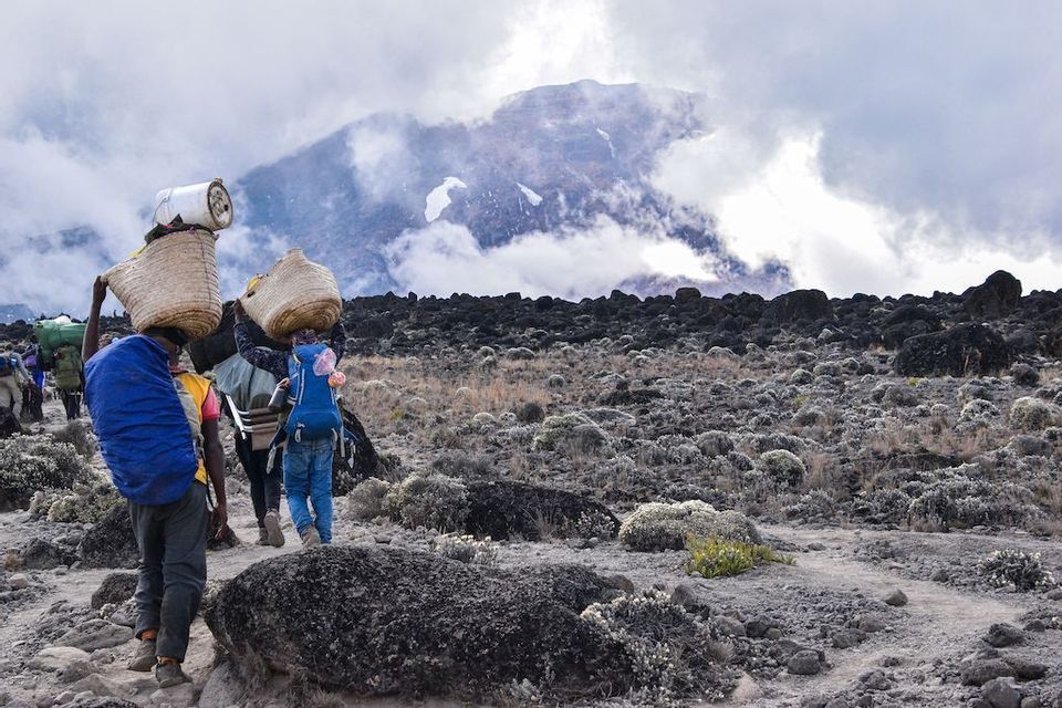 Un voyage de groupe WeRoad avec des porteurs transportant des paniers tressés marche sur un sentier rocheux vers un sommet de montagne couvert de nuages.