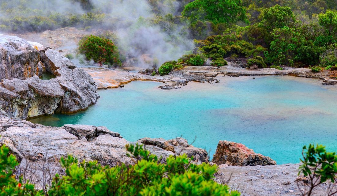 Ein leuchtend türkisblauer Geothermiepool, umgeben von grauen Felsen und grüner Vegetation, mit aufsteigendem Dampf im Hintergrund.