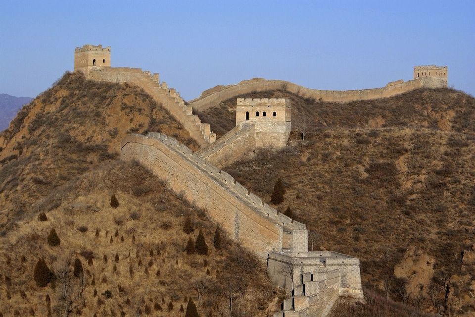 Una larga muralla de piedra fortificada con atalayas serpentea por colinas áridas y empinadas bajo un cielo azul claro.