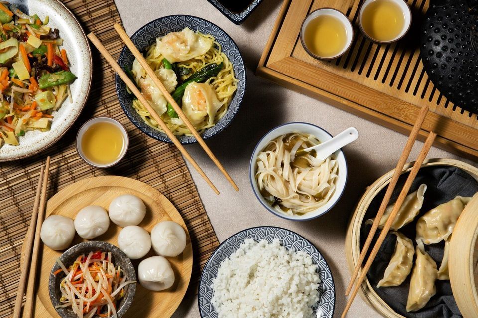 Una vista aérea de una comida asiática extendida sobre una mesa, que incluye fideos, empanadillas, arroz y té.