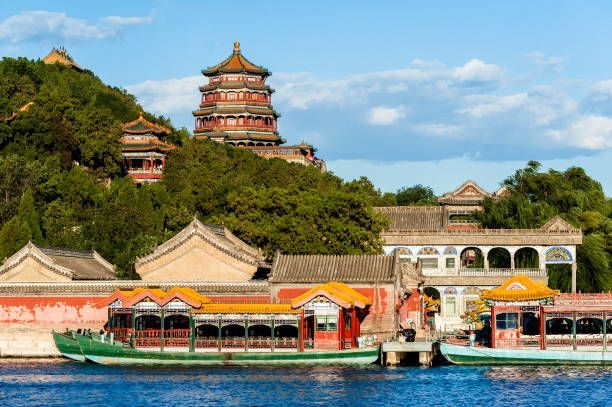 Barcos chinos tradicionales están atracados en un lago frente a una colina verde coronada por una pagoda histórica y otros edificios.