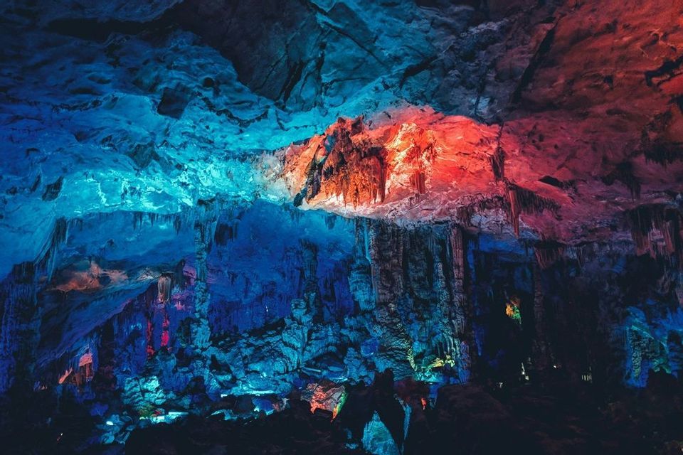 El interior de una gran cueva con estalactitas y formaciones rocosas iluminadas por luces azules y rojas brillantes.