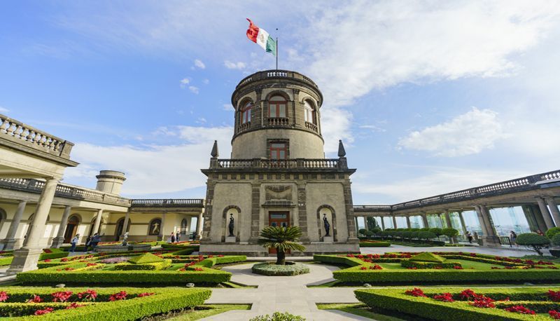 A stone castle with a central tower flying the Mexican flag, surrounded by a formal garden with manicured green hedges and flowers.