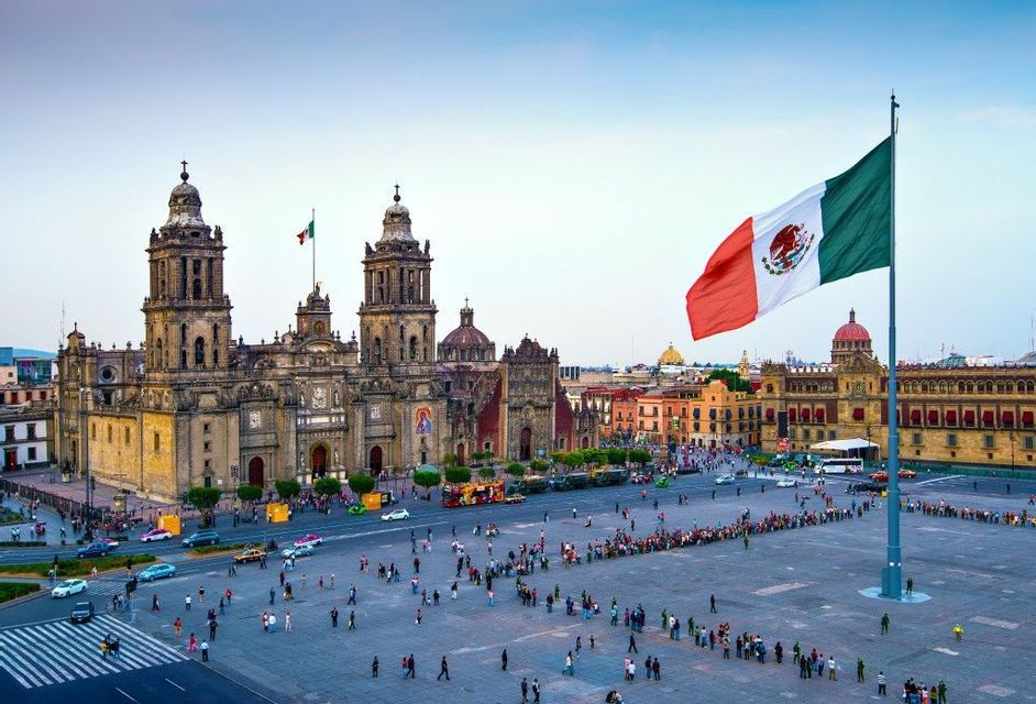 A large Mexican flag waves over a historic city square bustling with people, with an ornate cathedral in the background.